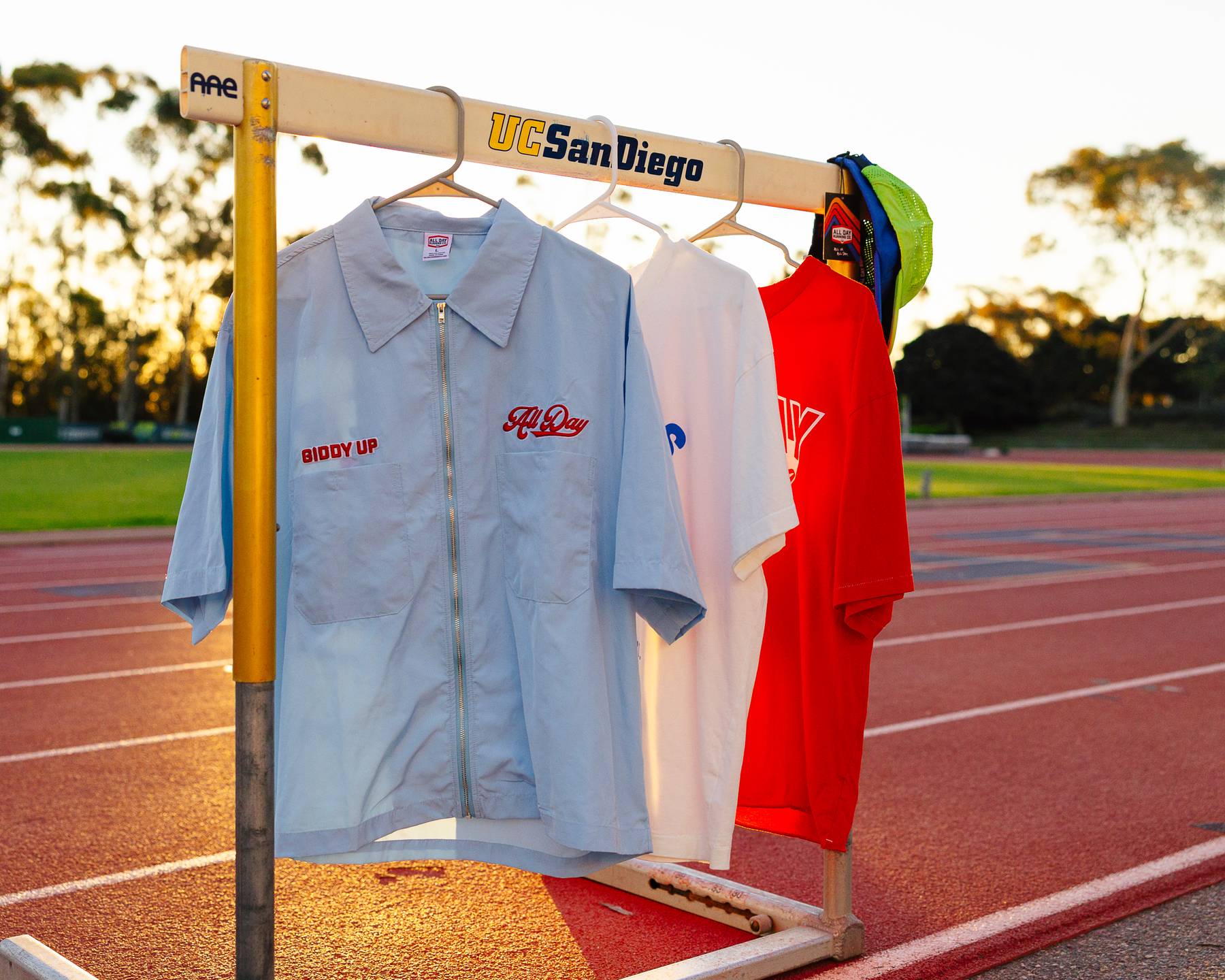 Three All Day Running Co shirts hanging on a hurdle on a track 